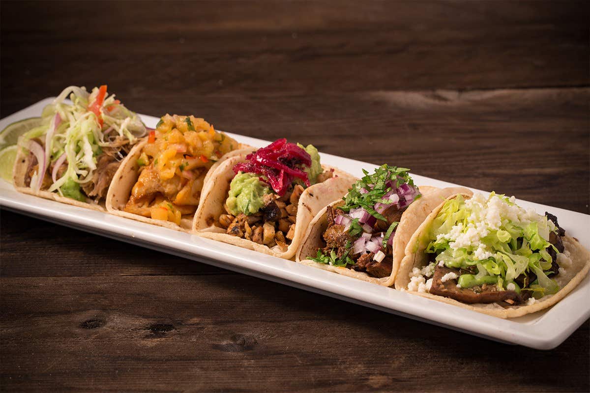 A long white plate with five assorted tacos, each topped with different colorful ingredients, on a wooden table.