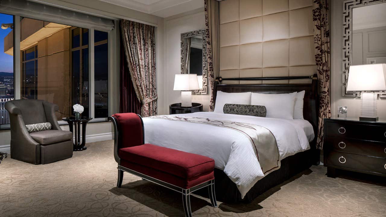 Luxurious hotel room with a large bed, nightstands, lamps, a red bench, an armchair, and a city view through the window.