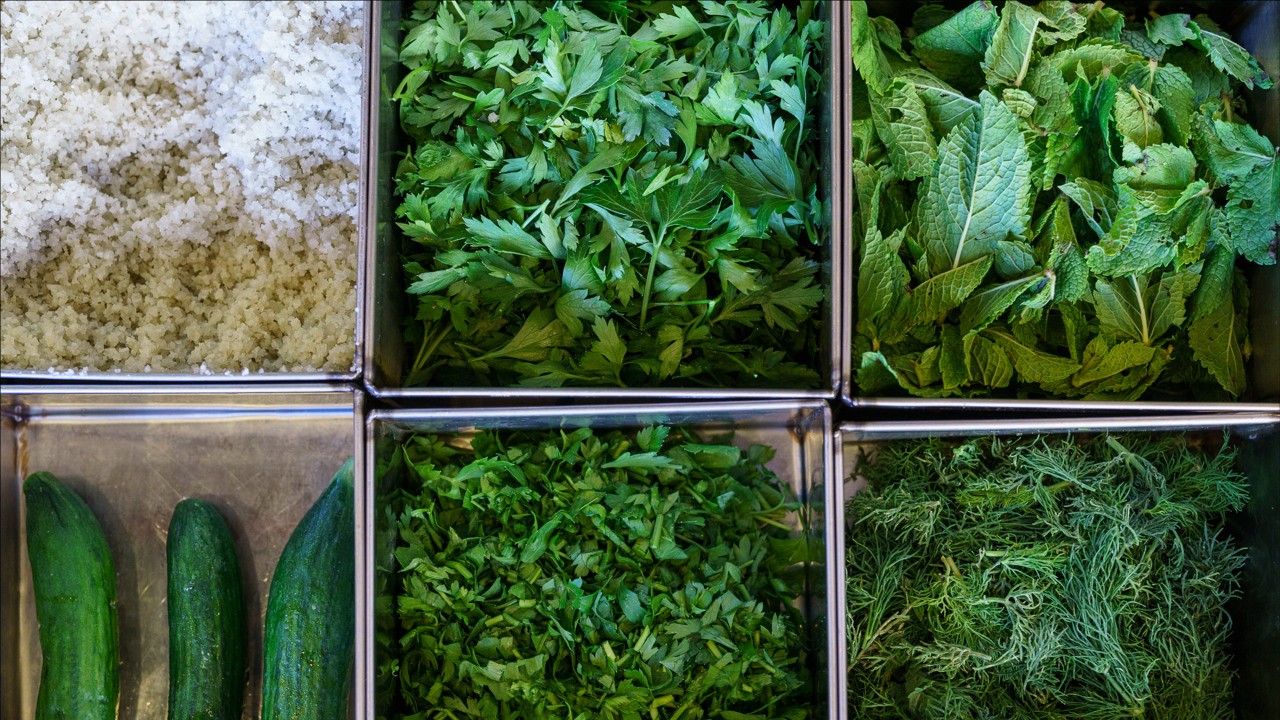 Assorted fresh herbs and vegetables, including cucumbers, parsley, mint, dill, and a box of coarse salt displayed in trays.