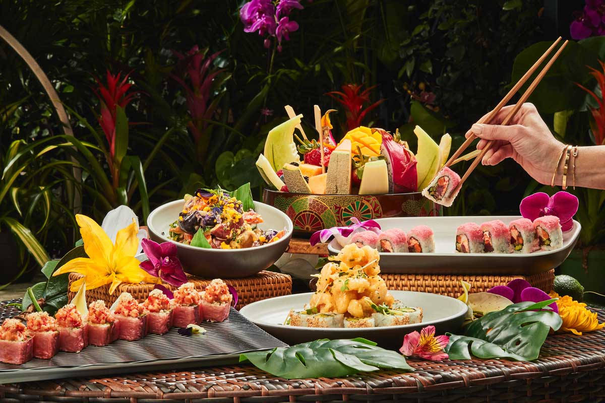 An assortment of colorful sushi dishes and fresh fruits on a table, with a hand using chopsticks to pick up a sushi roll.