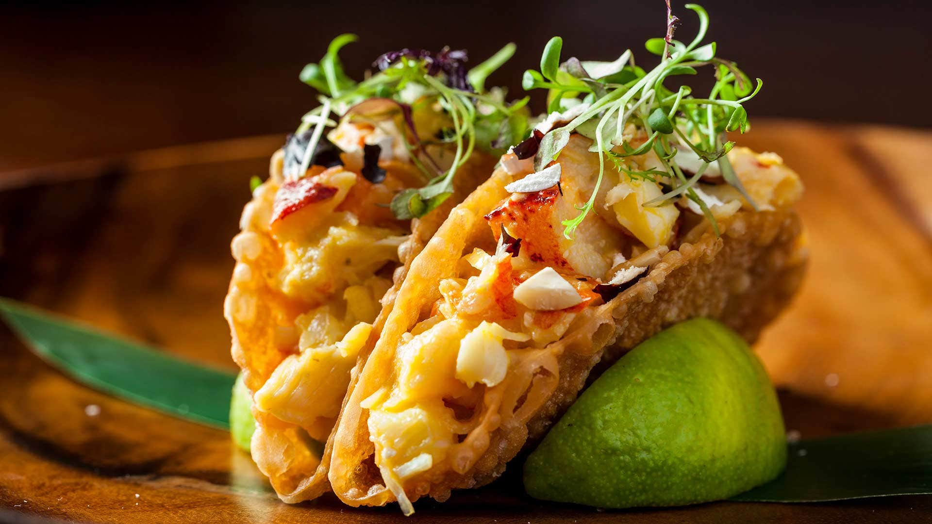 Close-up of two gourmet tacos filled with lobster and topped with fresh greens, garnished with a lime wedge.