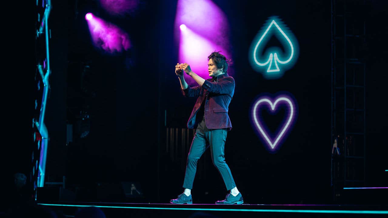 Shin Lim on stage with purple and blue lighting, holding an object with neon spade and heart symbols in the background.