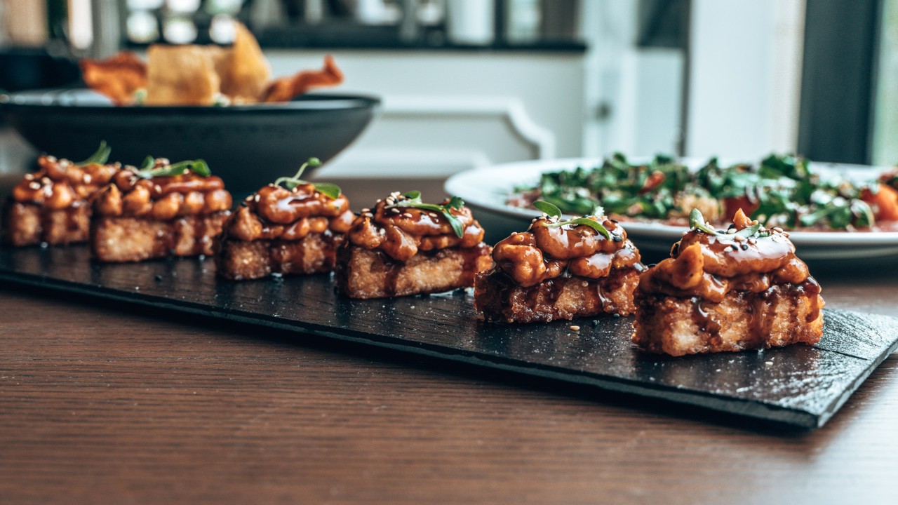 A row of delicious bite-sized appetizers on a black slate, with a salad and a bowl of chips in the background.