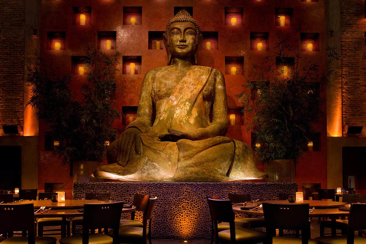 A large Buddha statue in a dimly lit restaurant surrounded by candlelit tables and warm, ambient lighting.