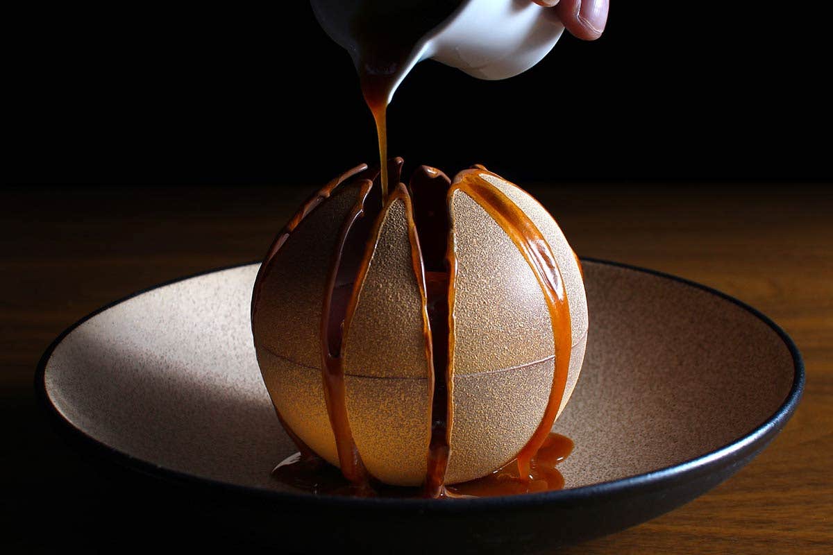 A caramel sauce is being poured over a golden, sphere-shaped dessert on a plate.