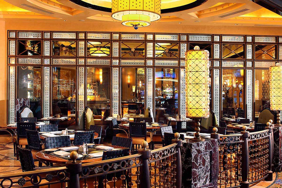 Elegant restaurant interior with ornate decor, warm lighting, wooden tables, and cushioned chairs, featuring large glass windows.