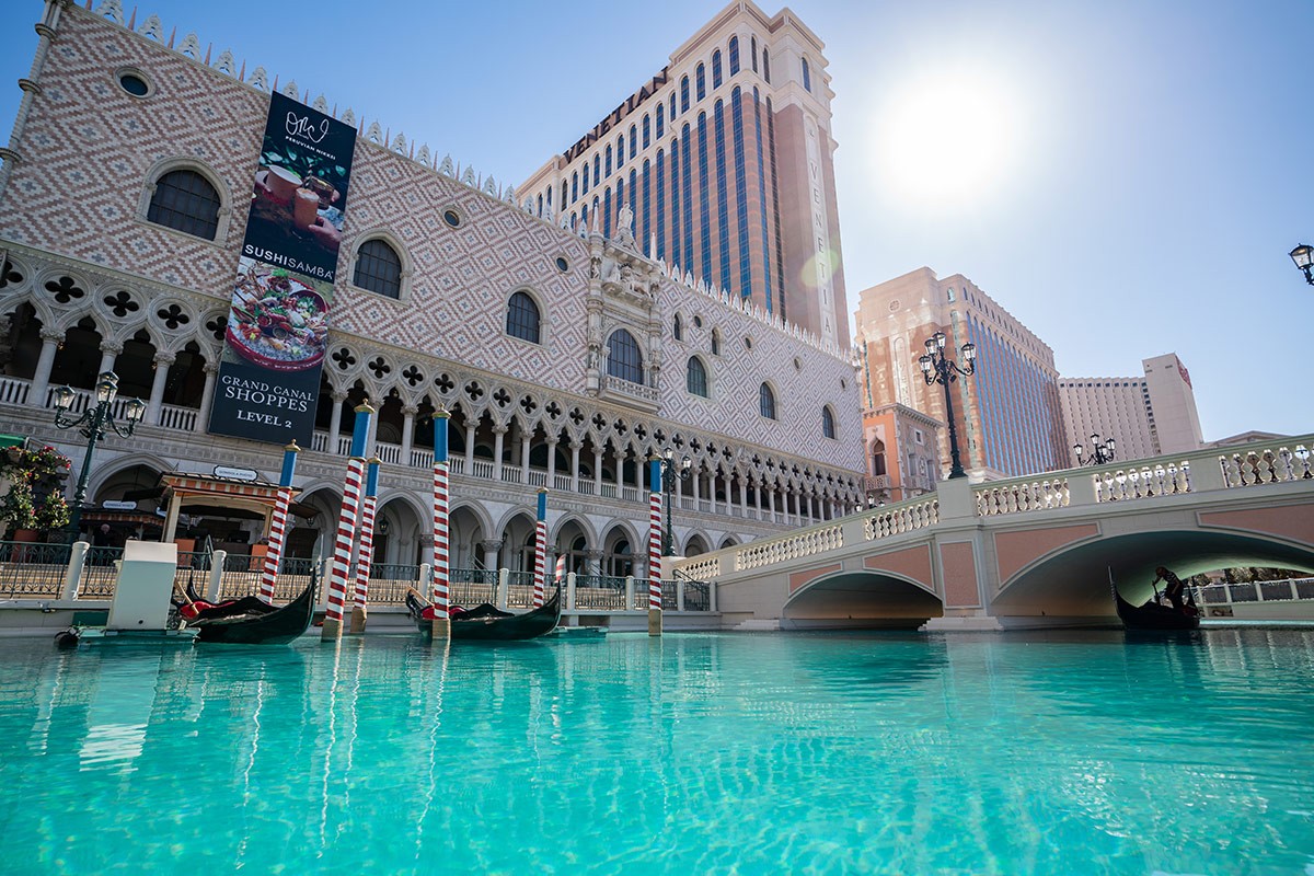 A turquoise canal at a Venetian-themed hotel and casino with gondolas, under a clear blue sky and bright sun.