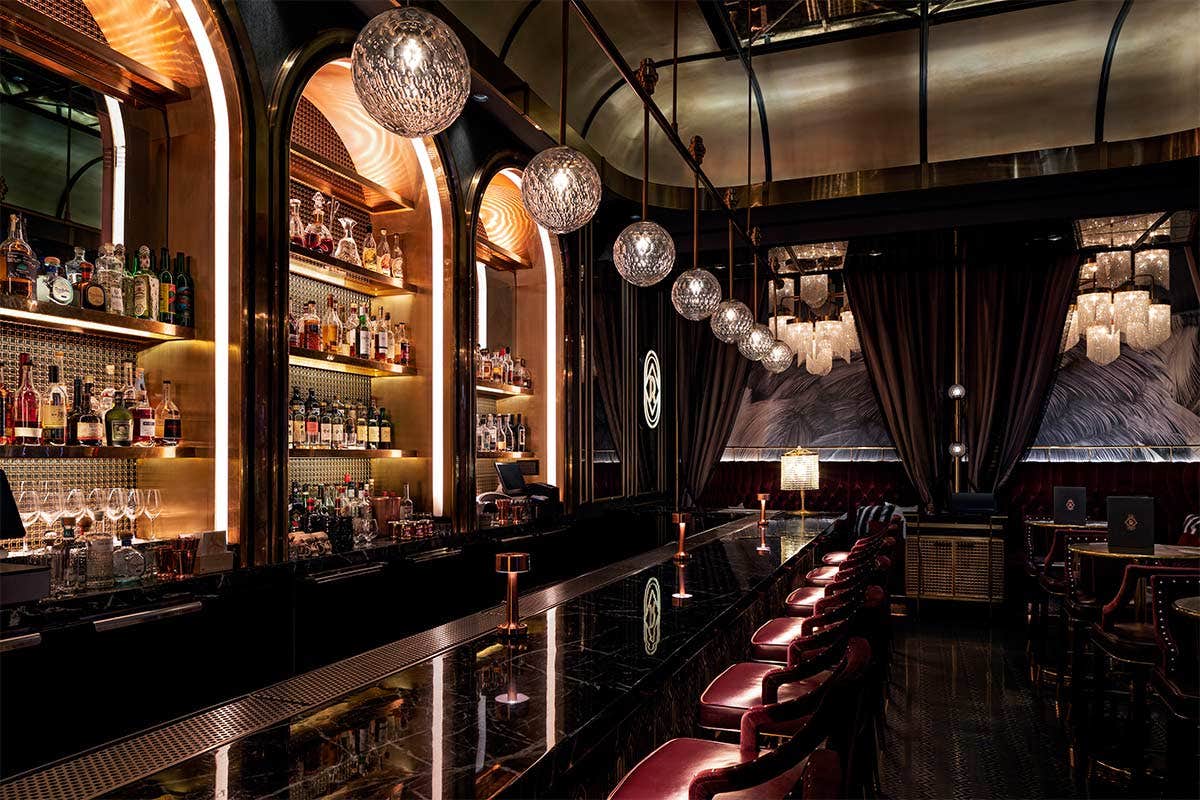 Dimly lit upscale bar with crystal globe lights, backlit shelves of bottles, dark wood paneling, and leather bar stools.