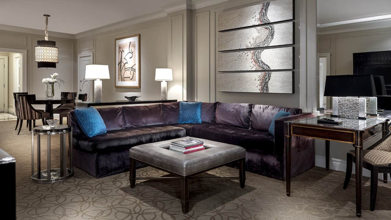 Modern living room with a plush sectional sofa, blue cushions, an ottoman, wall art, table lamps, and adjoining dining area.