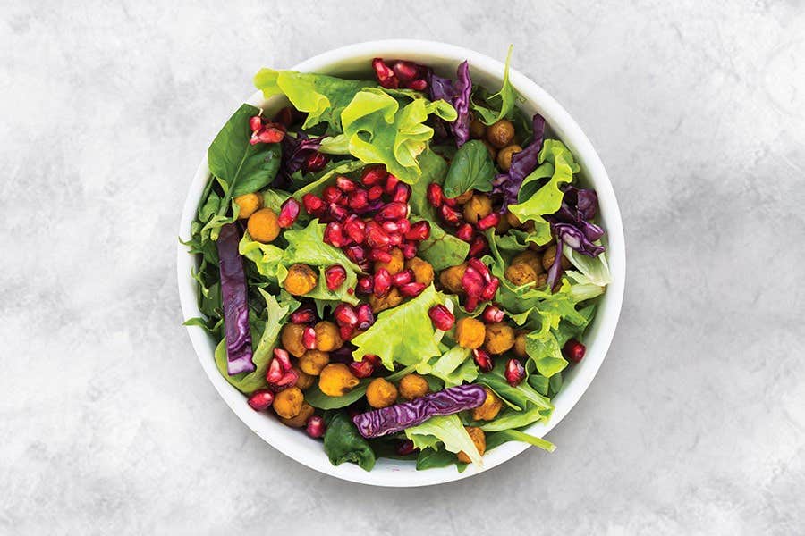 A vibrant salad with mixed greens, chickpeas, red cabbage, and pomegranate seeds in a white bowl against a gray background.