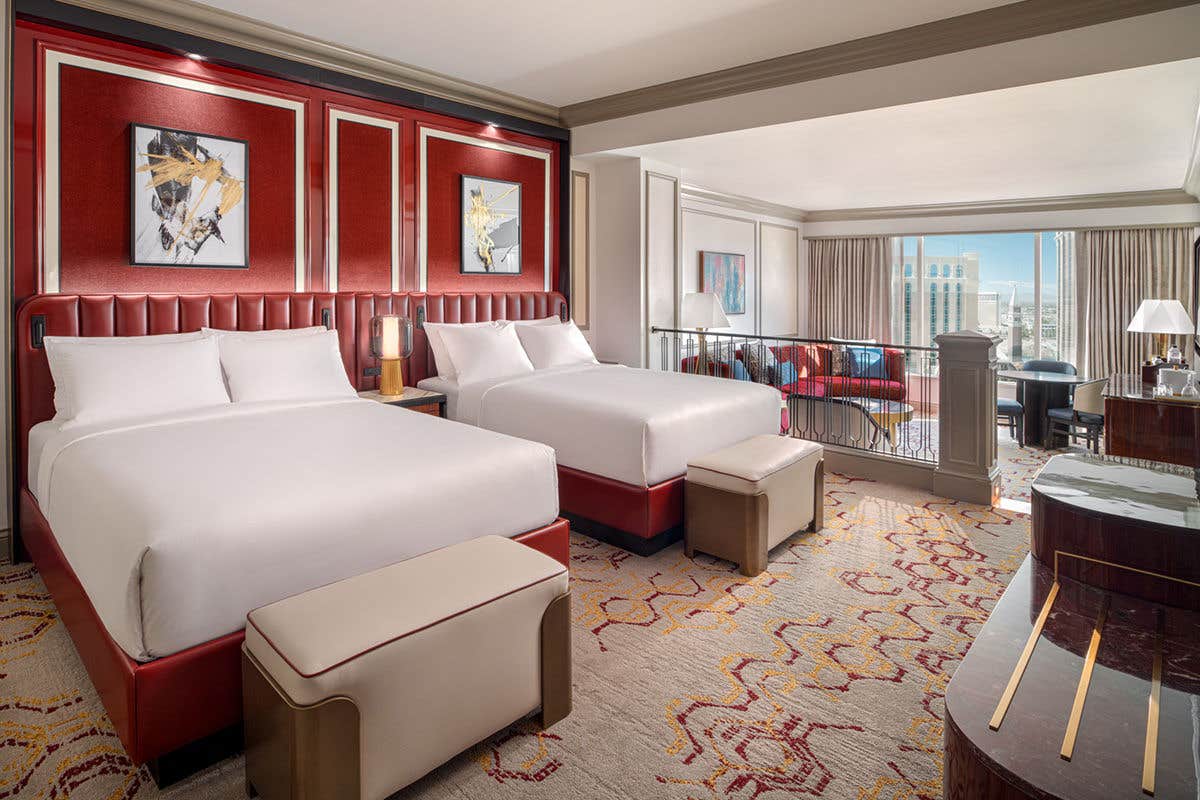 A suite in The Venetian South Tower showing two queen beds in the foreground with a red wall behind them, and the living space in the back with a view of the city