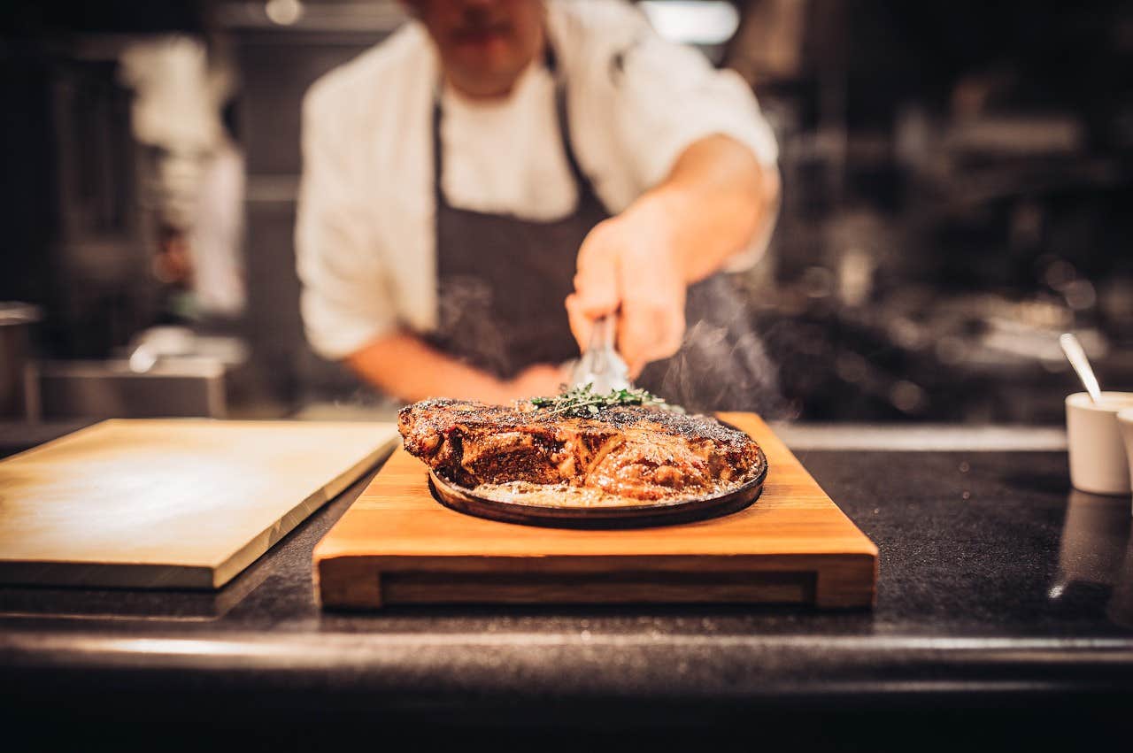 Chef garnishing a cooked steak with herbs on a wooden board in a professional kitchen.