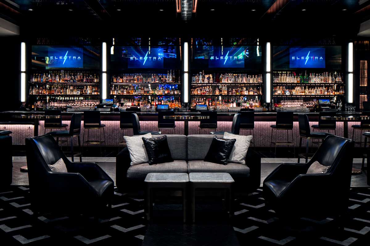 A modern lounge with a well-lit bar, leather chairs, and a gray sofa in the center, displaying "Elettra" signs above the bar.