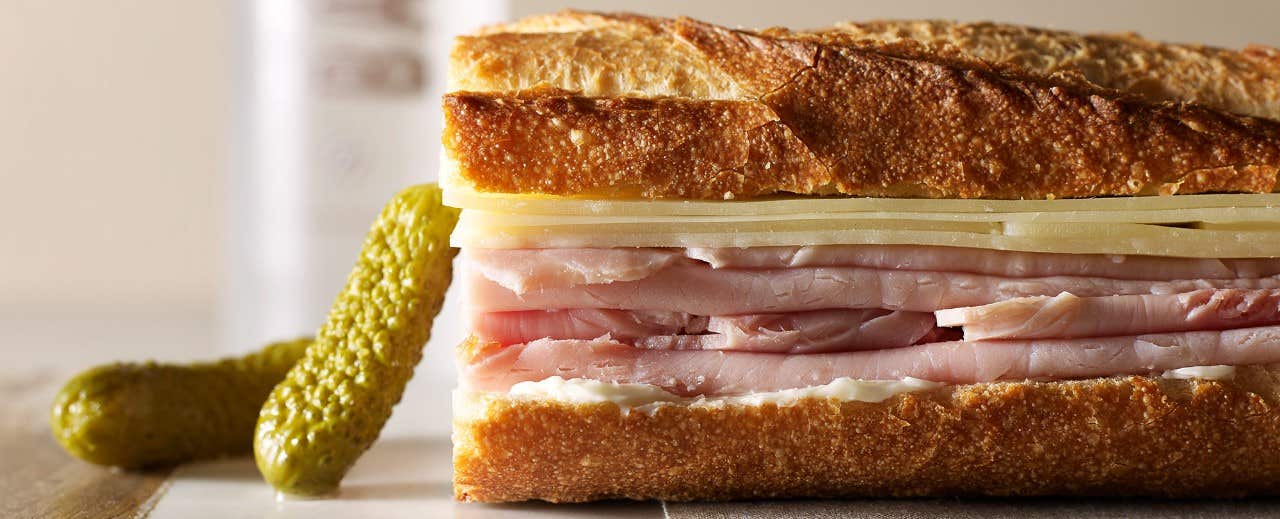 A close-up of a ham and cheese baguette with two pickles on the side.