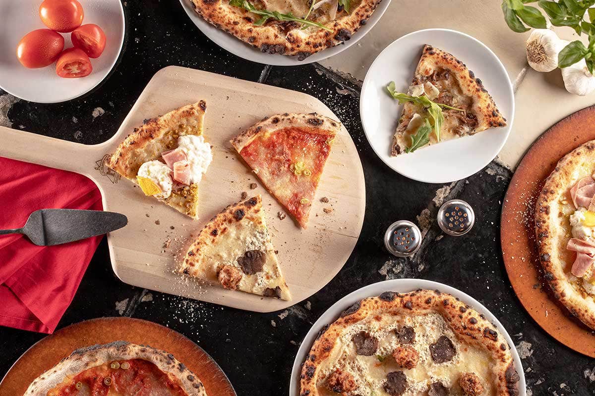 Assorted pizzas on a table; a pizza peel holding three slices, surrounded by whole pies, tomatoes, garlic, and a red napkin.