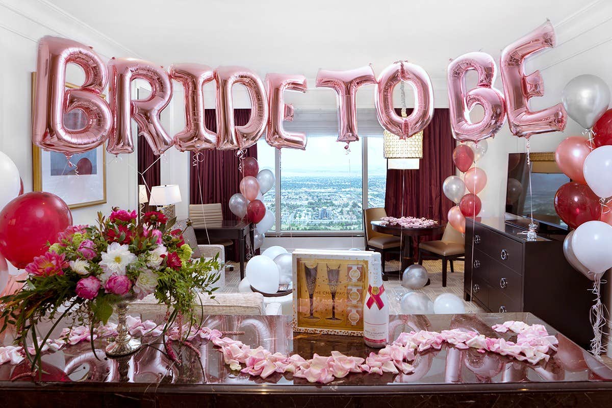 Hotel room decorated with "BRIDE TO BE" balloons, flowers, and a view of a cityscape through a large window.