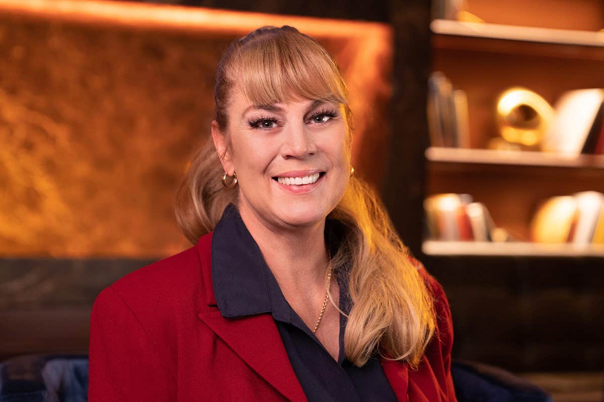 Smiling woman in a red blazer with blonde hair, sitting in a warmly lit room with bookshelves in the background.