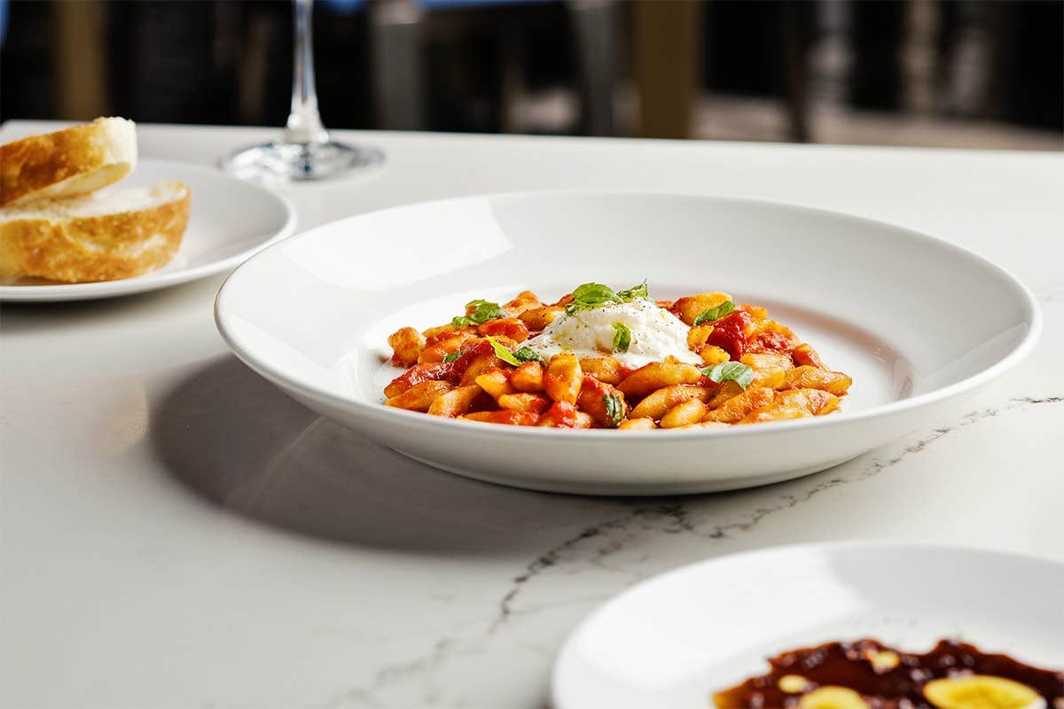 A white bowl of pasta with tomato sauce and herbs on a marble table with bread and wine in the background.