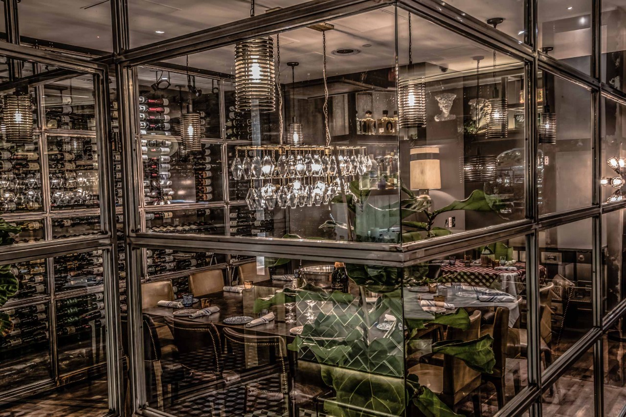 Elegant glass-enclosed dining room with modern lighting, lush plants, and tables set for a meal.