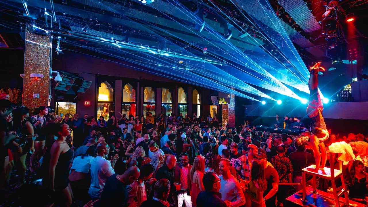 Crowded nightclub with a performer doing a handstand on a stool and vibrant laser lights illuminating the dance floor.