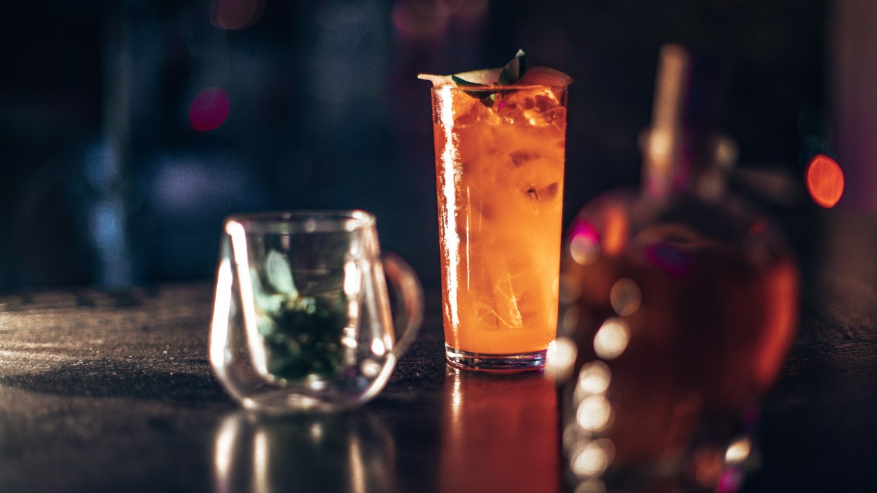 Tall glass of amber cocktail garnished with an orange peel on a dimly lit bar with a glass mug and bottle in the foreground.