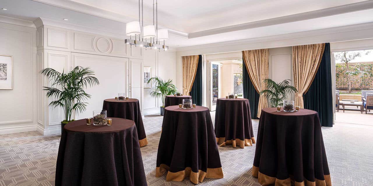 An elegant Hospitality Suite with round tables covered in dark brown tablecloths, plants, and framed art, featuring large windows and a chandelier.