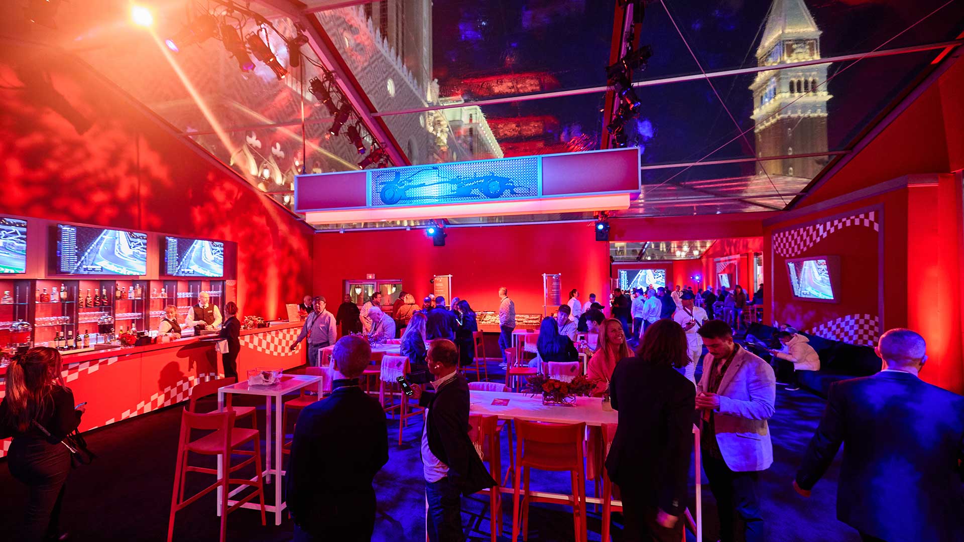 A lively party scene under a glass roof, with people mingling, red decor, and screen displays.