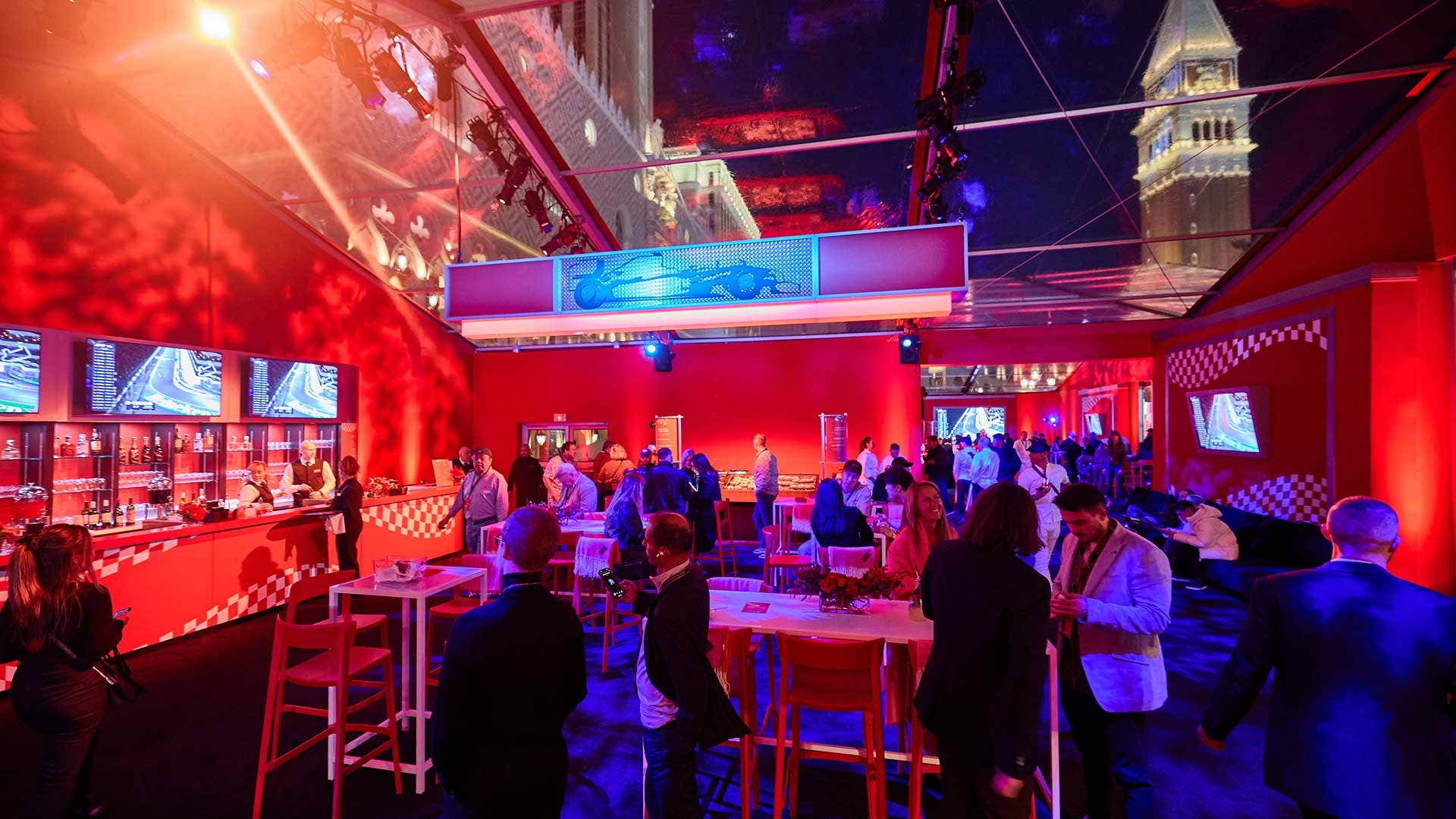 A lively party scene under a glass roof, with people mingling, red decor, and screen displays.