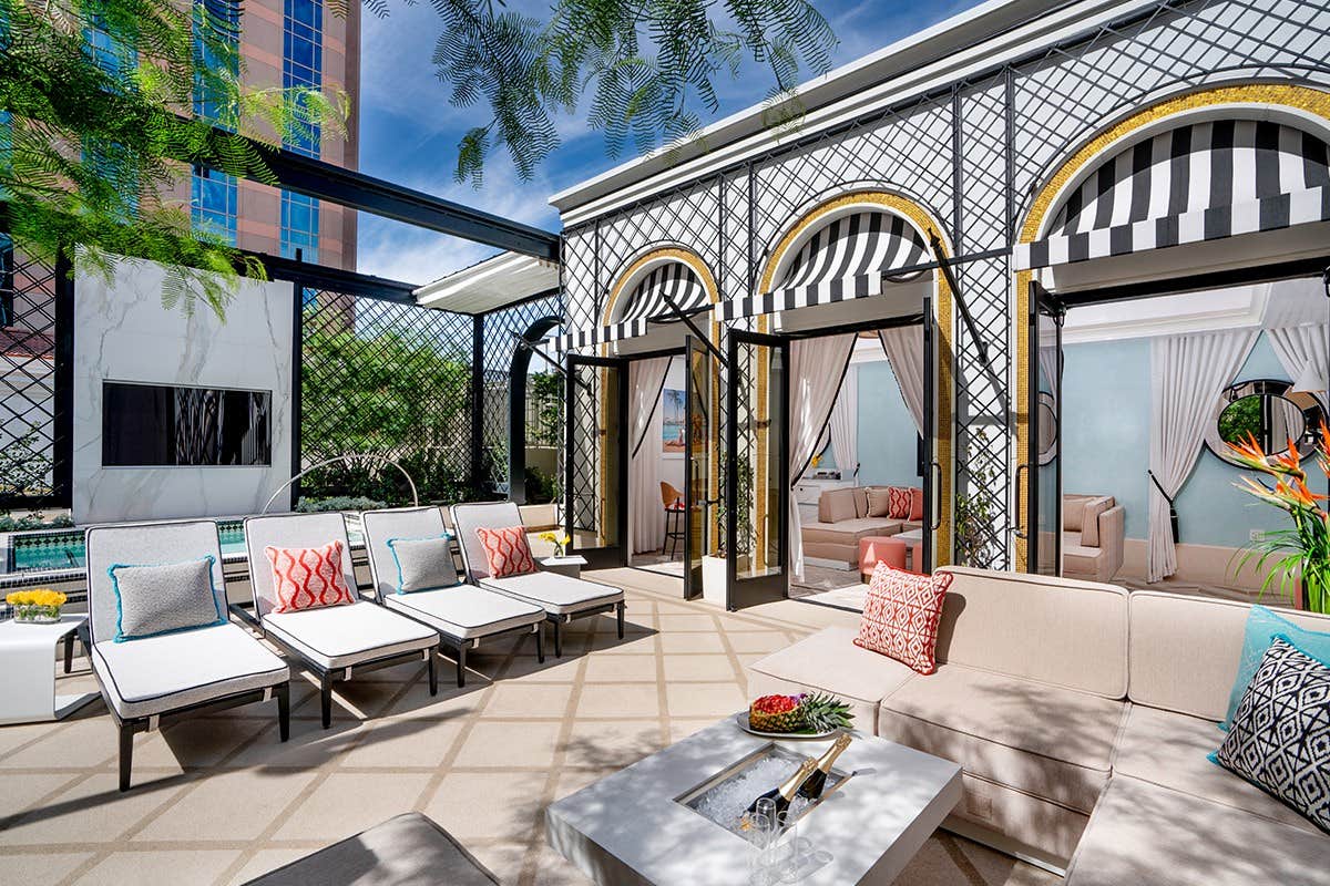 Modern outdoor patio with lounge chairs, a sofa, and patterned pillows. Open doors lead to a chic indoor area.