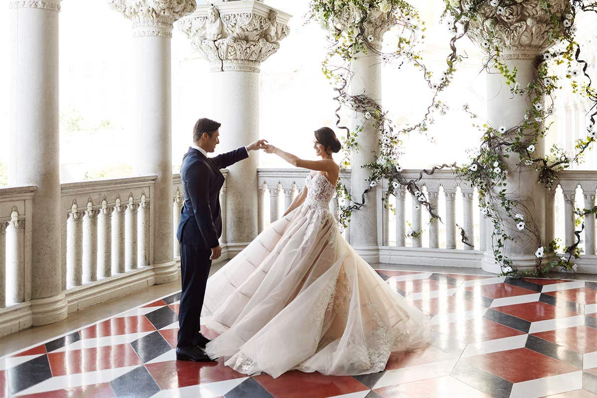 A couple dances in elegant wedding attire on a patterned floor in a columned venue adorned with greenery and flowers.