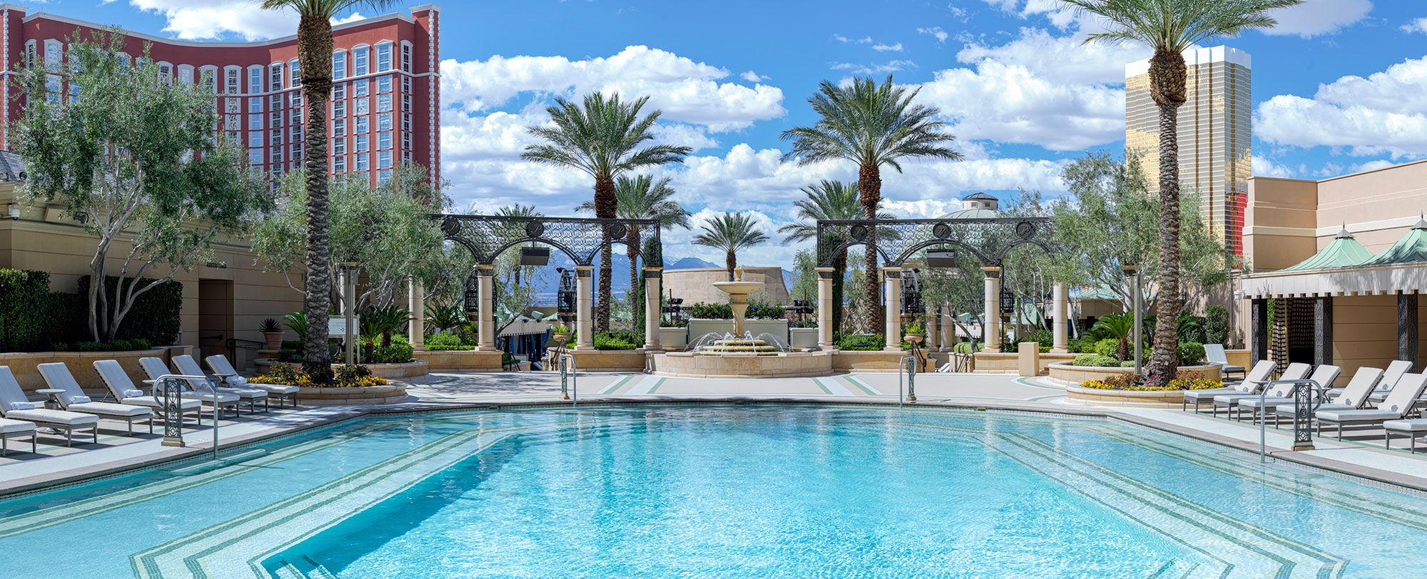 A luxurious swimming pool with lounge chairs, palm trees, and a lavish resort building in the background on a sunny day.