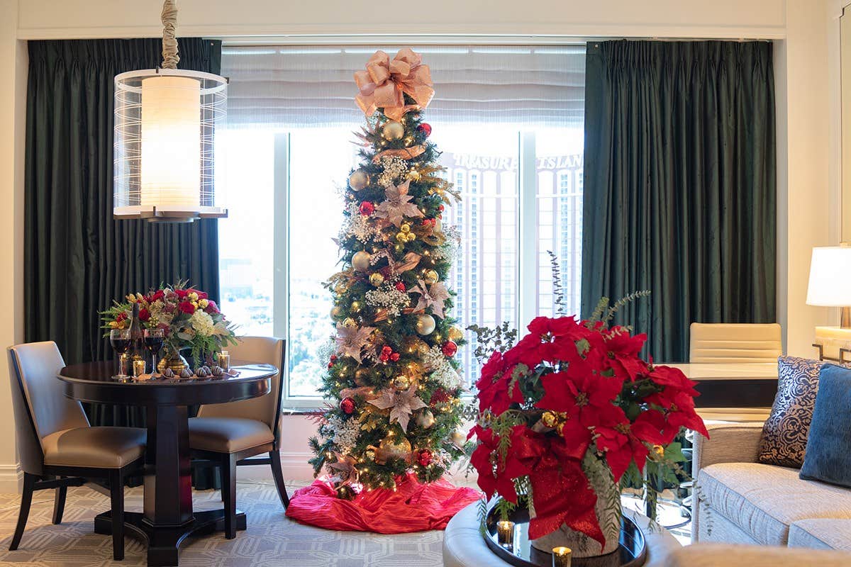 A decorated Christmas tree in a living room with poinsettias, a round table with flowers, and large windows with city view.