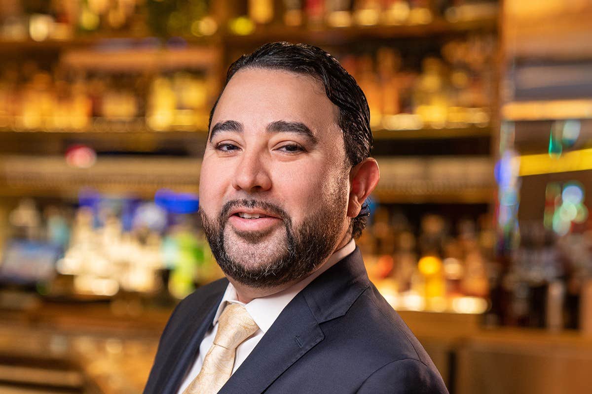 A man with a beard, wearing a suit and tie, smiles in a warmly lit environment like a bar or restaurant.