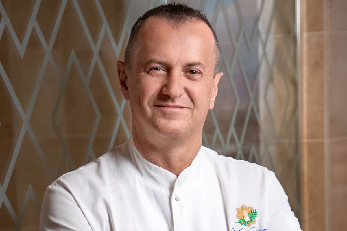 A smiling chef in a white coat stands with arms crossed against a patterned glass background.