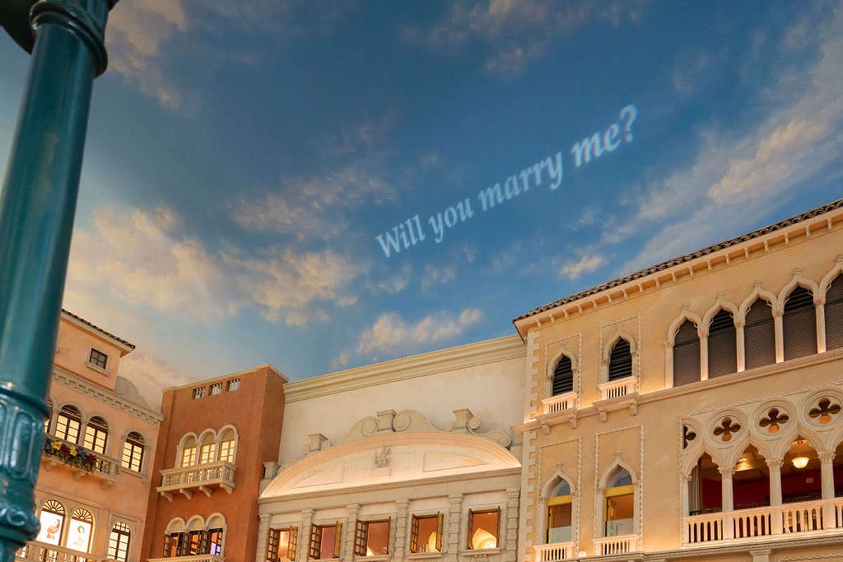 Proposal message "Will you marry me?" written in the sky above a European-style building facade.