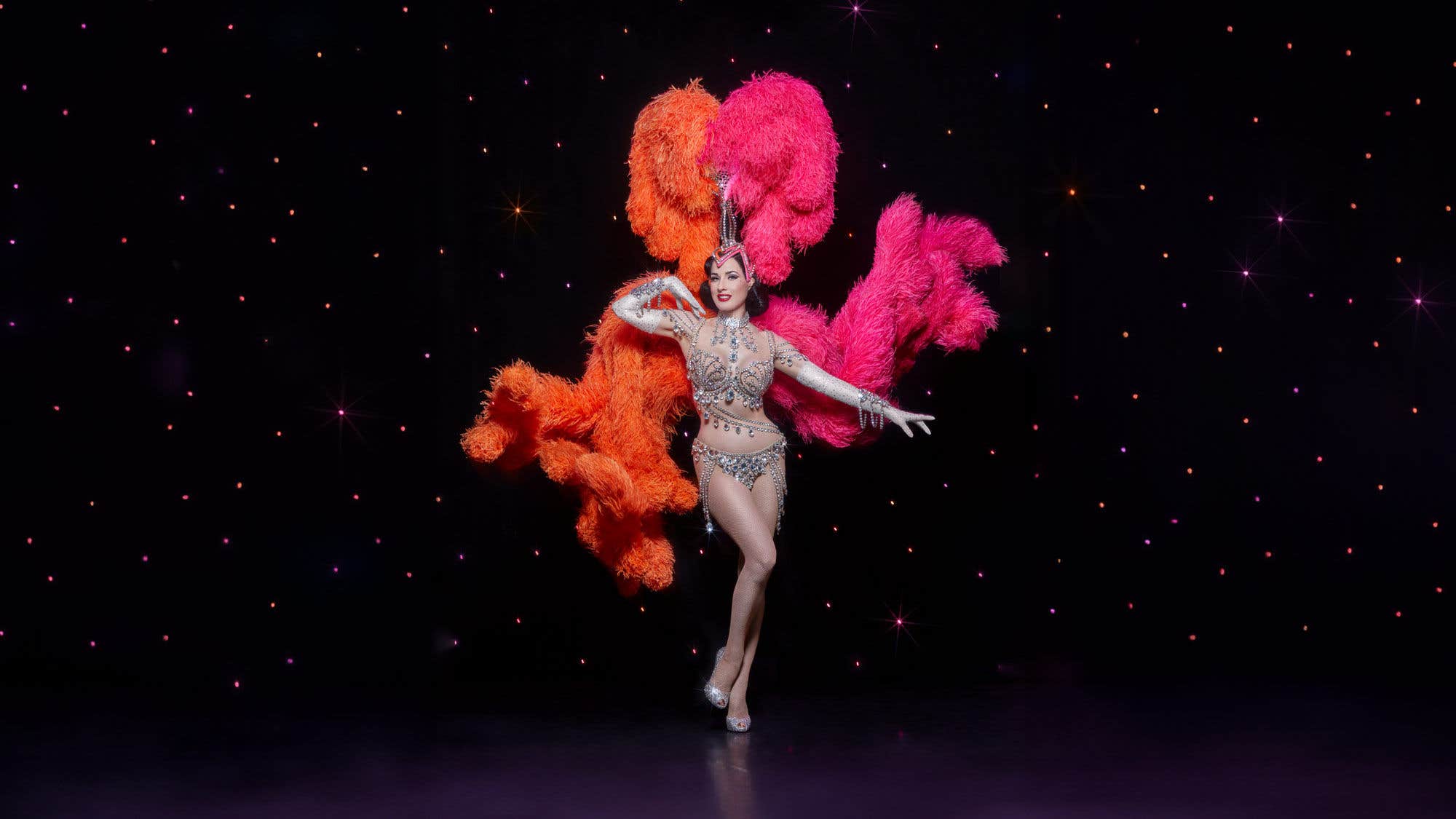 Dita Von Teese in a diamond-covered outfit with an orange and pink headdress in front of a starry backdrop