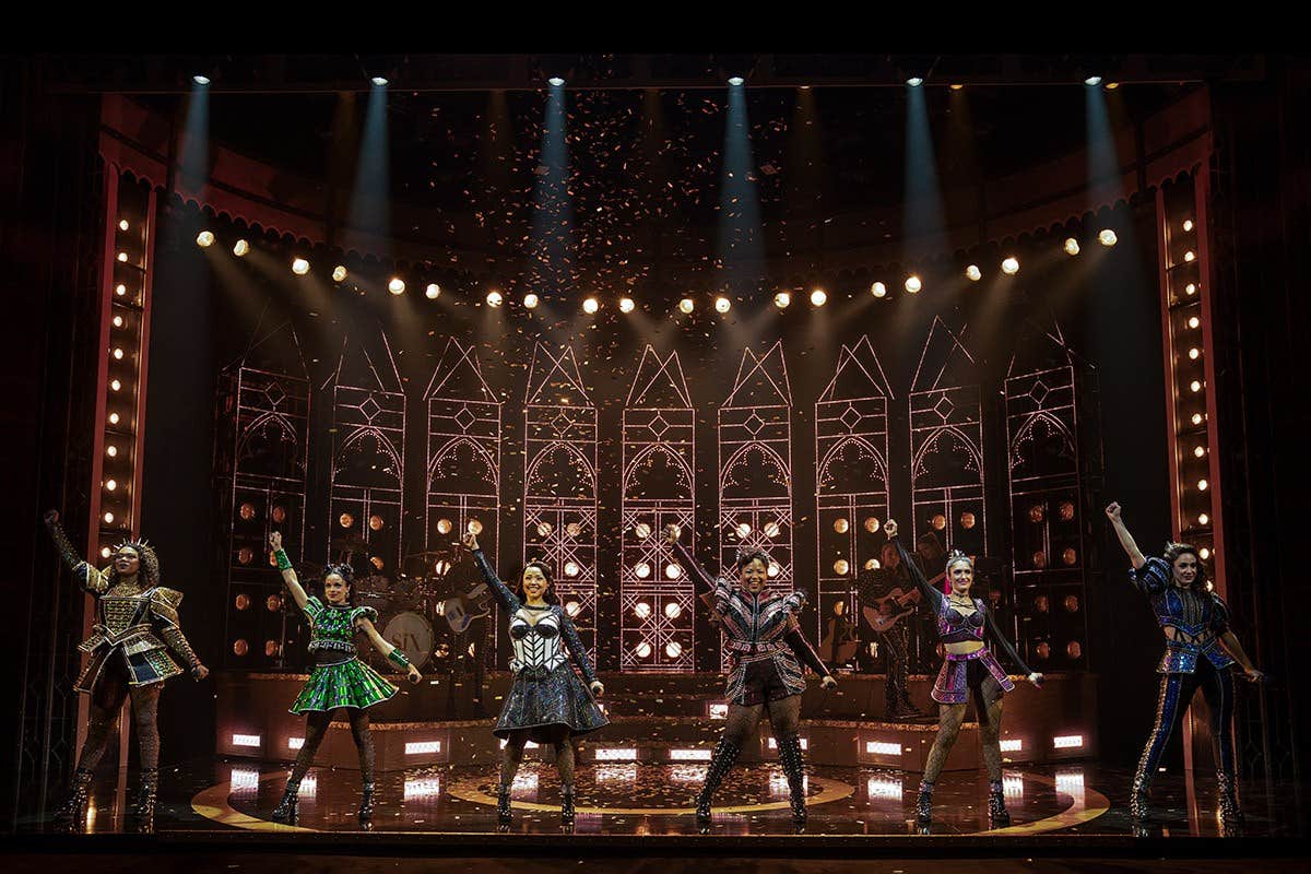 Six performers in colorful, ornate costumes on a stage with a flashy, medieval backdrop, raising their arms triumphantly.