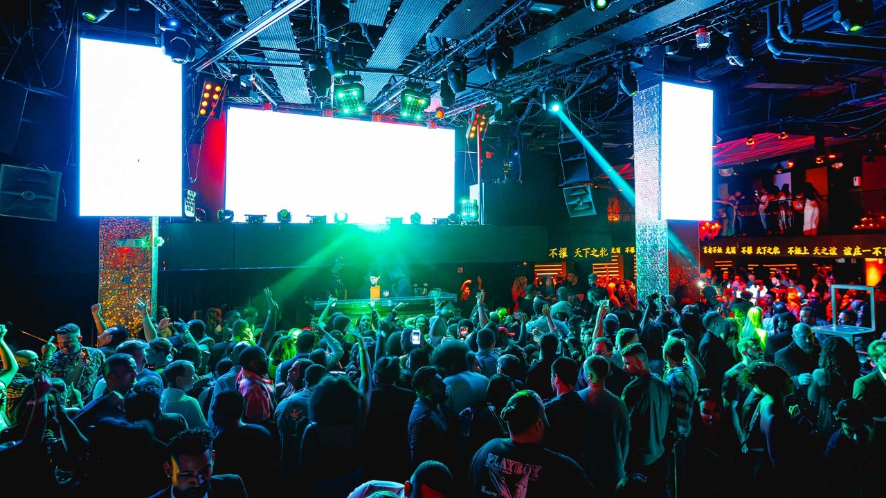 A crowded nightclub with people dancing, neon lights, and a DJ performing on a stage with large screens in the background.