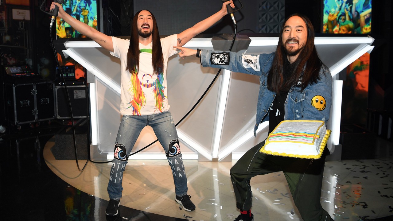 Two men with long hair and beards, one holding a microphone and the other holding a cake, in front of a DJ booth.