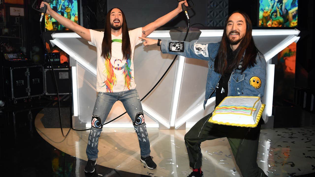 Two men with long hair and beards, one holding a microphone and the other holding a cake, in front of a DJ booth.