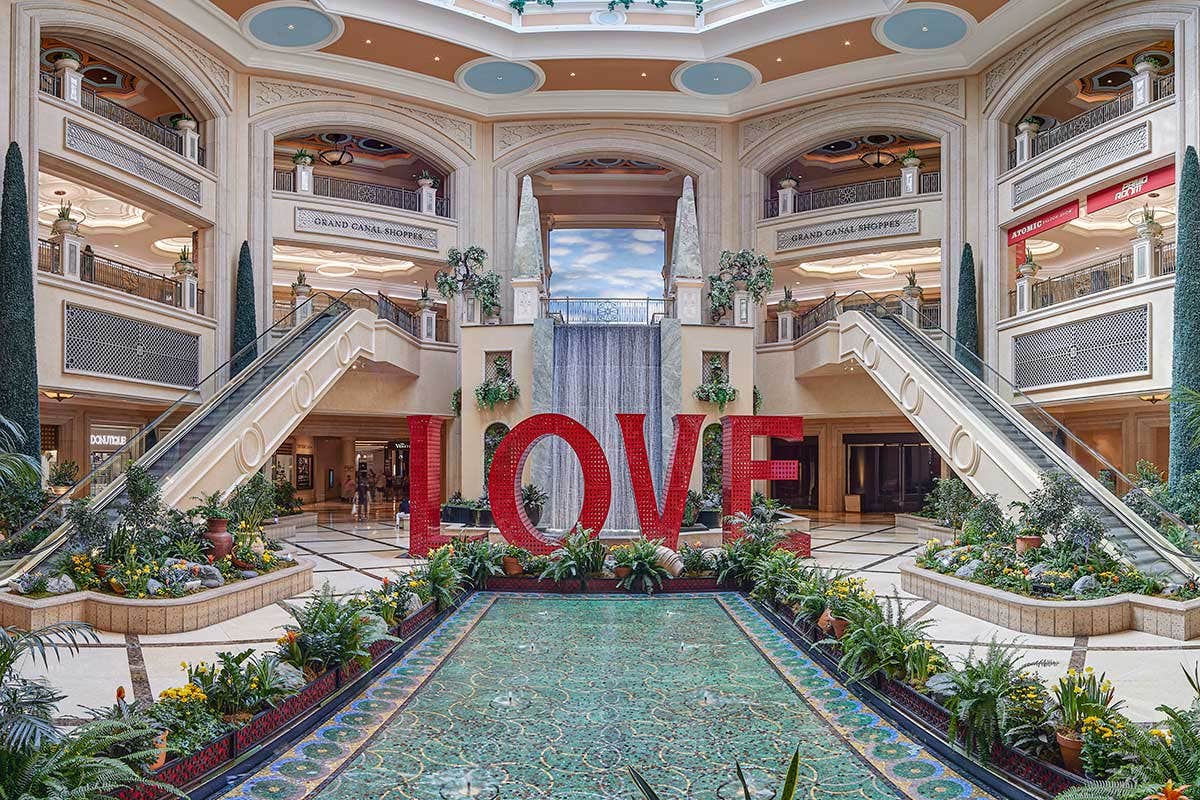 A shopping mall with a large red "LOVE" sculpture, greenery, and a fountain under a domed skylight.