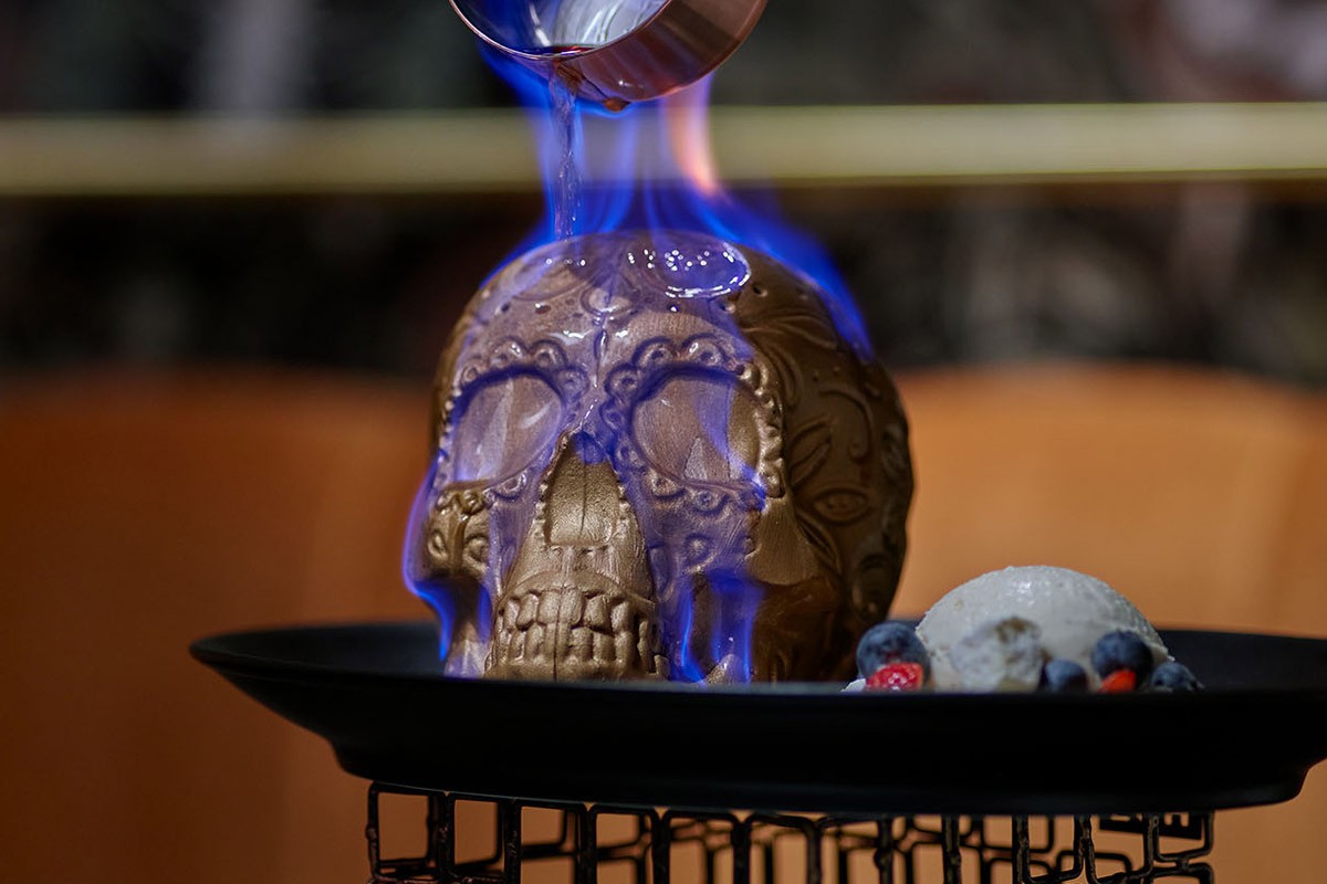 A flaming skull-shaped dessert on a plate with a scoop of ice cream and berries.