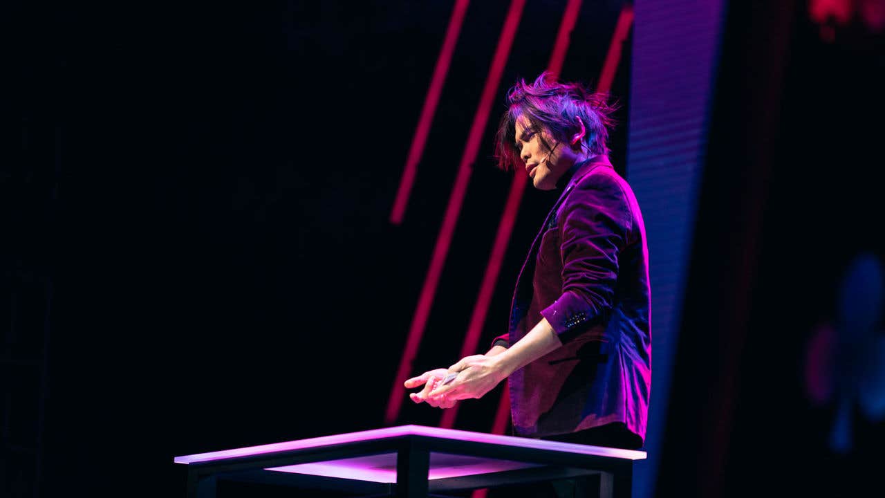 Shin Lim on stage illuminated by pink and purple lights, behind a table, with a backdrop of pink light strips.