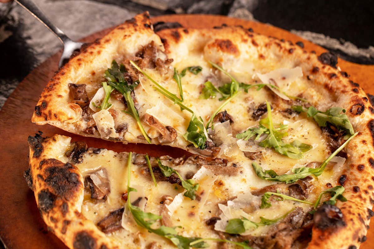 Freshly baked pizza topped with melted cheese, arugula, mushrooms, and shaved Parmesan, with one slice being lifted.