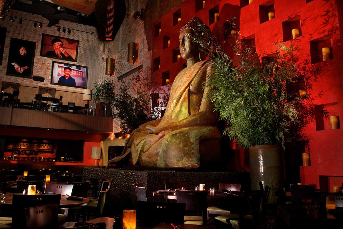 A large Buddha statue in a dimly lit restaurant with red walls and candles, greenery surrounding the statue.