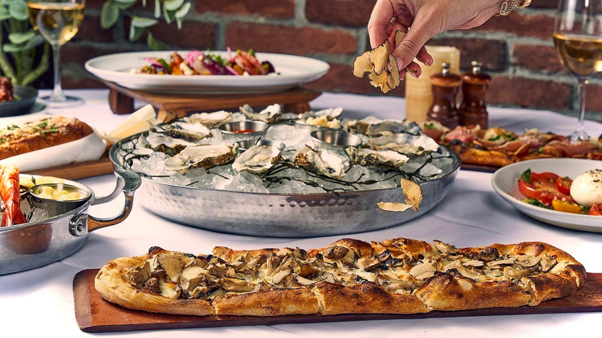 A hand sprinkling toppings over a dish in a seafood restaurant with oysters, flatbreads, and other dishes on the table.