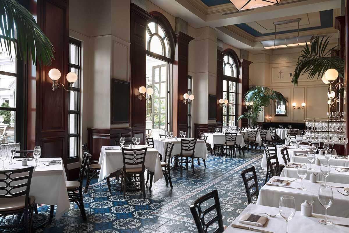 Elegant restaurant dining area with white tablecloths, dark wood paneling, large arched windows, and blue patterned floor tiles.