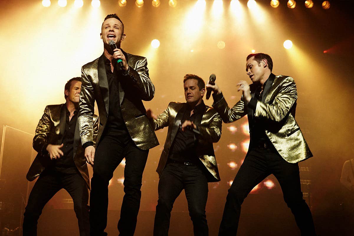 Four men in gold jackets perform energetically on stage with microphones under bright lights.