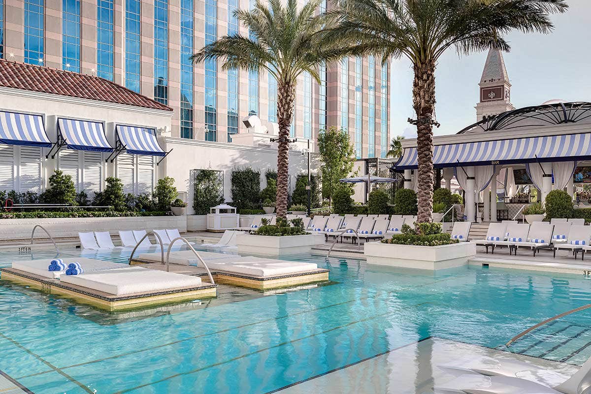 Luxurious outdoor pool area with lounge chairs, parasols, palm trees, and a nearby building equipped with blue-striped awnings.