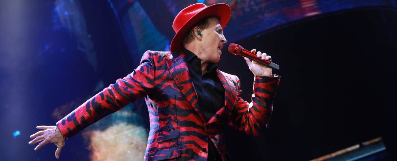 A person wearing a red hat and a red and black patterned suit sings into a red microphone on stage.